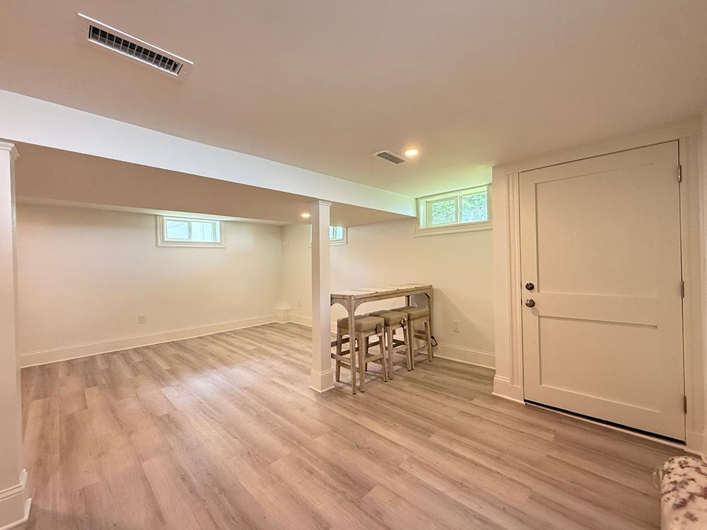 Empty, freshly renovated basement with pale wood-look floors, white walls, and a closed door.