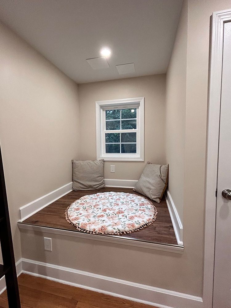 Reading nook with a window, cushions, and a round rug on a wooden platform.