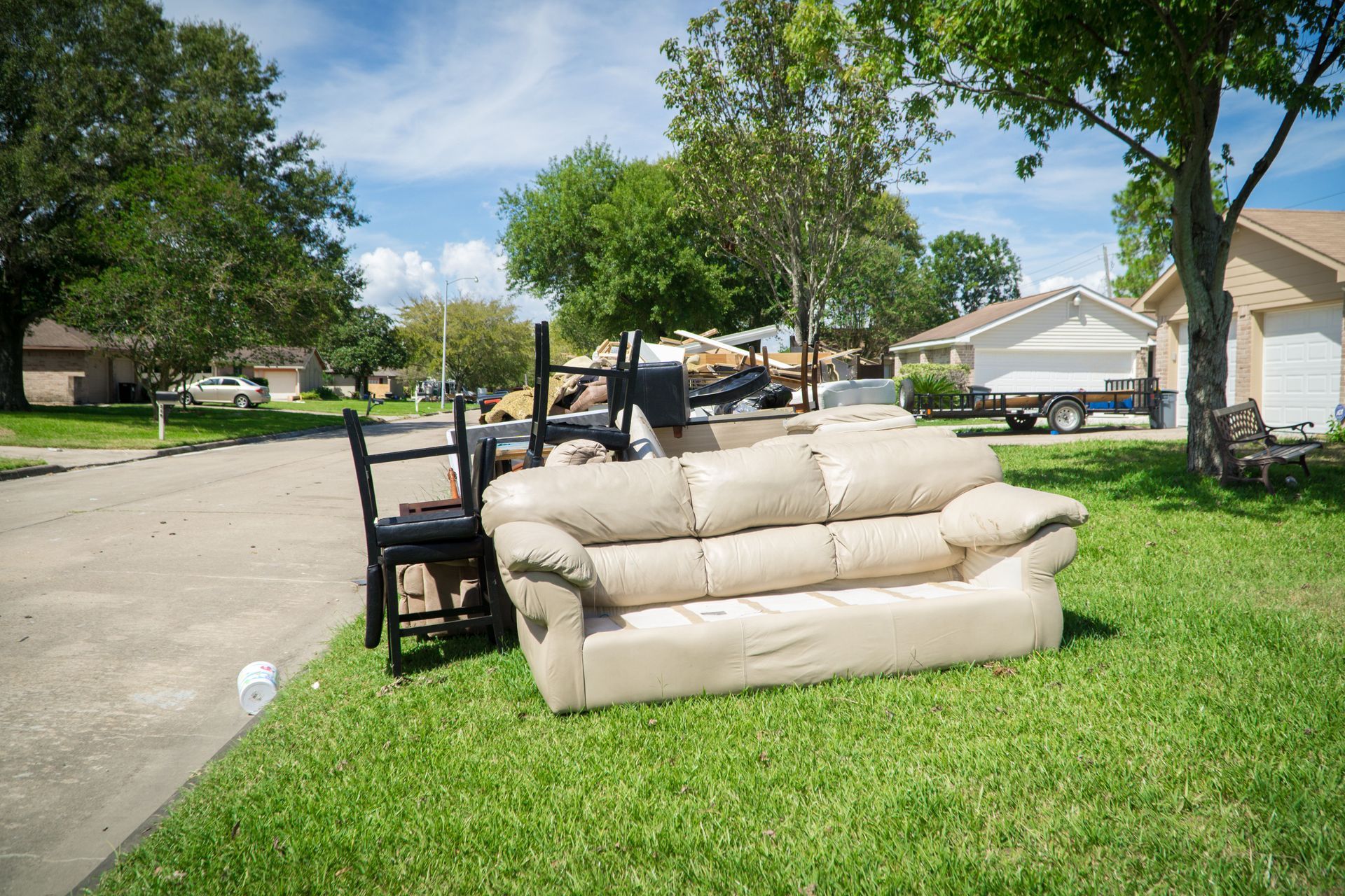 Curb-side view of flooded home debris: beige couch, chairs, and trash bags on grass in a residential neighborhood.