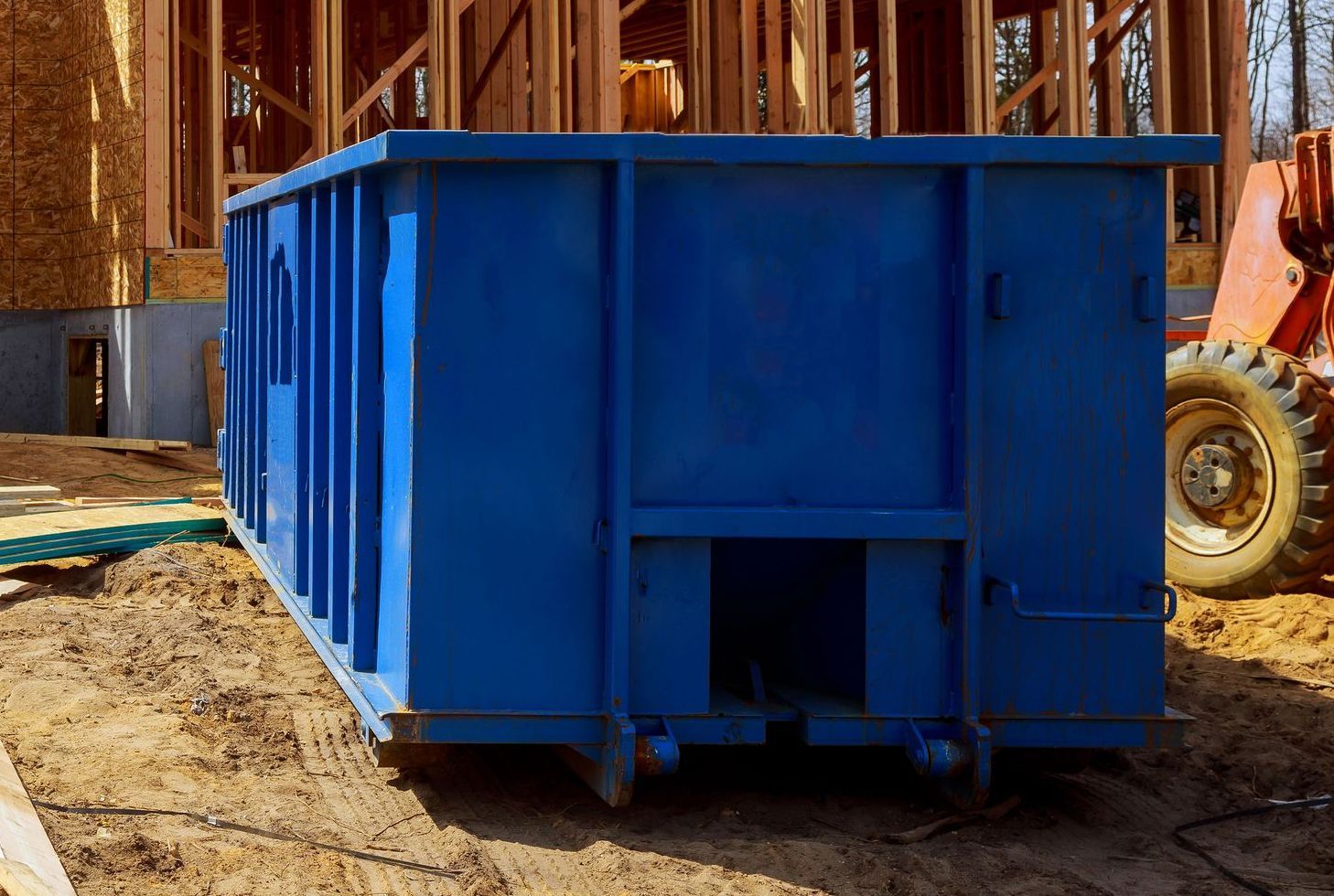Blue dumpster at a construction site with a partially built wooden structure in the background.