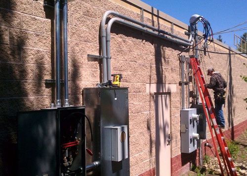 A man on a ladder is working on an electrical system on the side of a building.