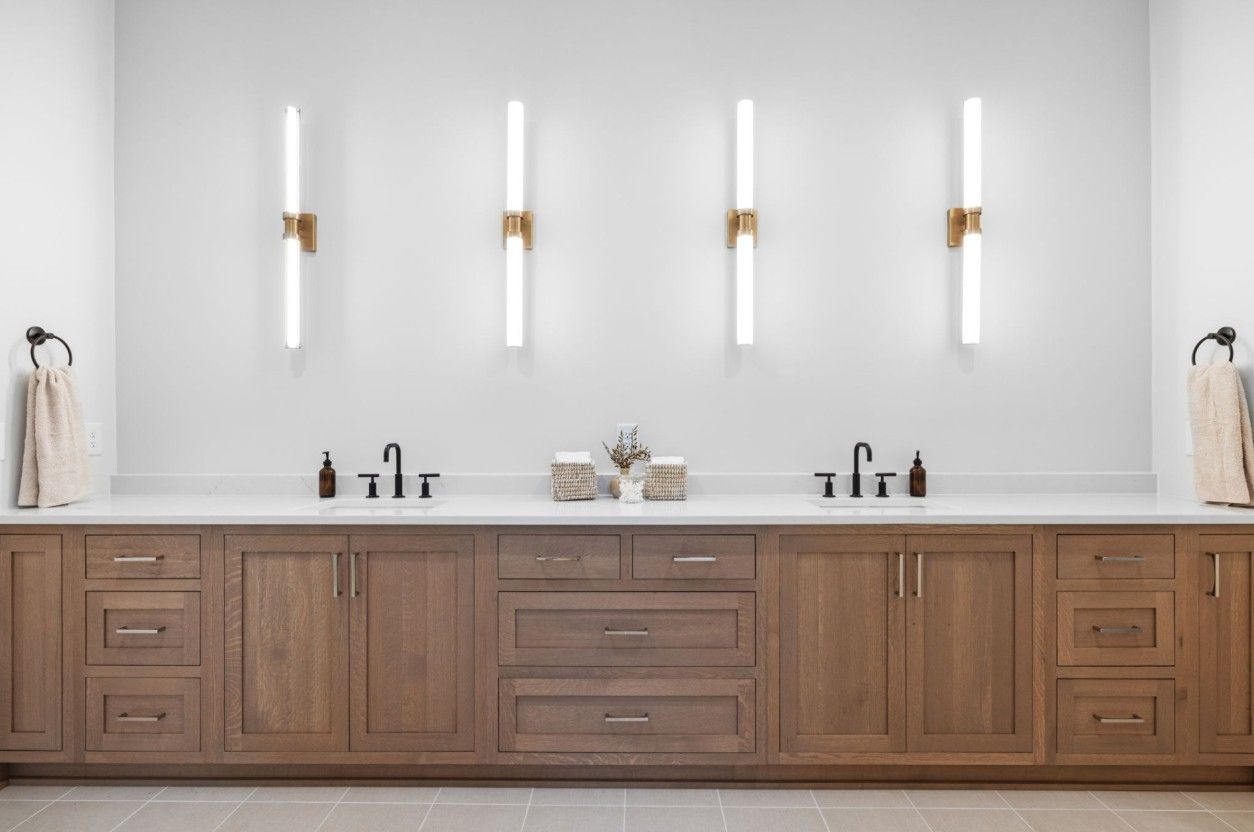 A double vanity with a wooden cabinet and white countertop, featuring four wall sconces and two faucets on a white wall.