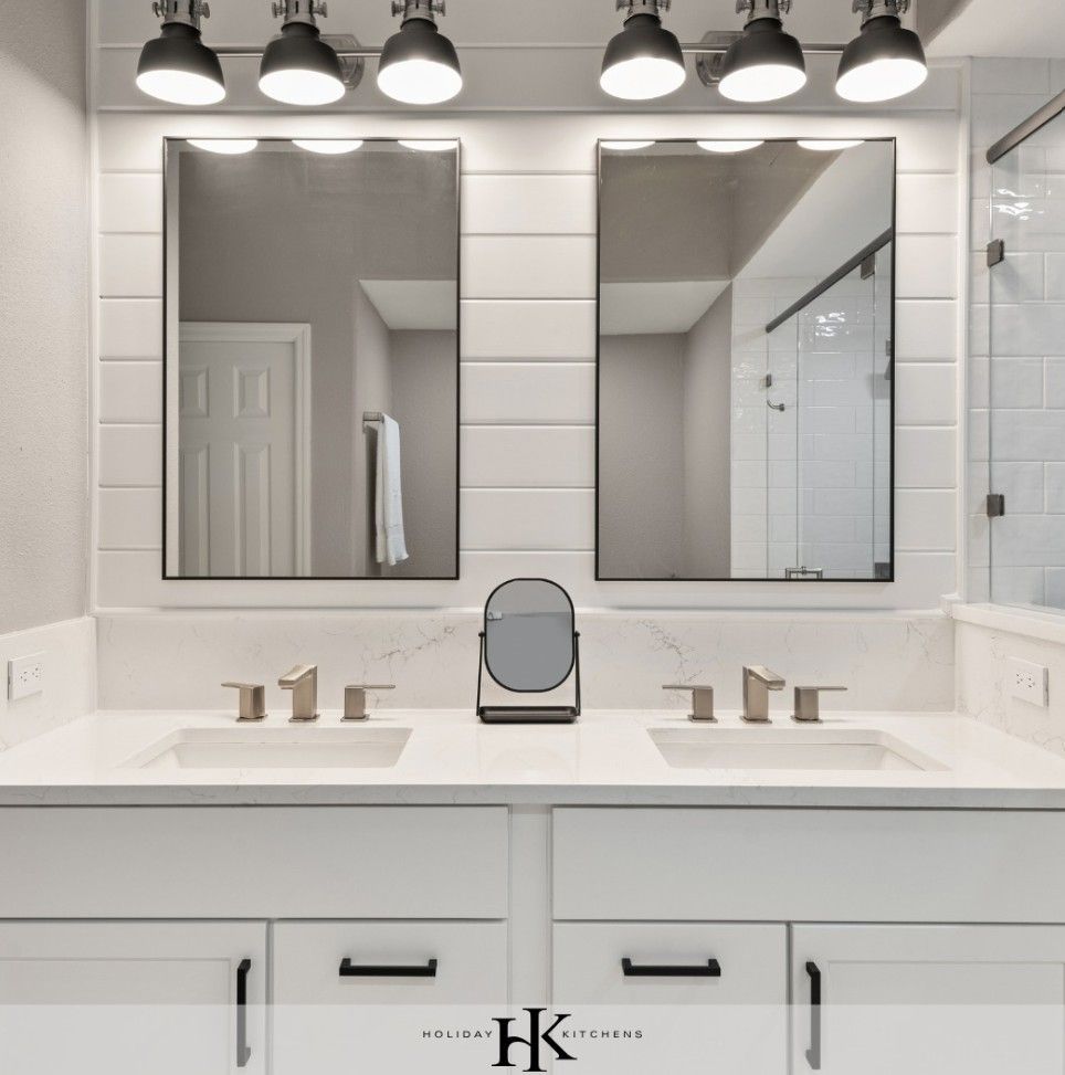 A bright modern bathroom featuring white cabinets, dual sinks, marble countertops, two mirrors, and overhead lighting.