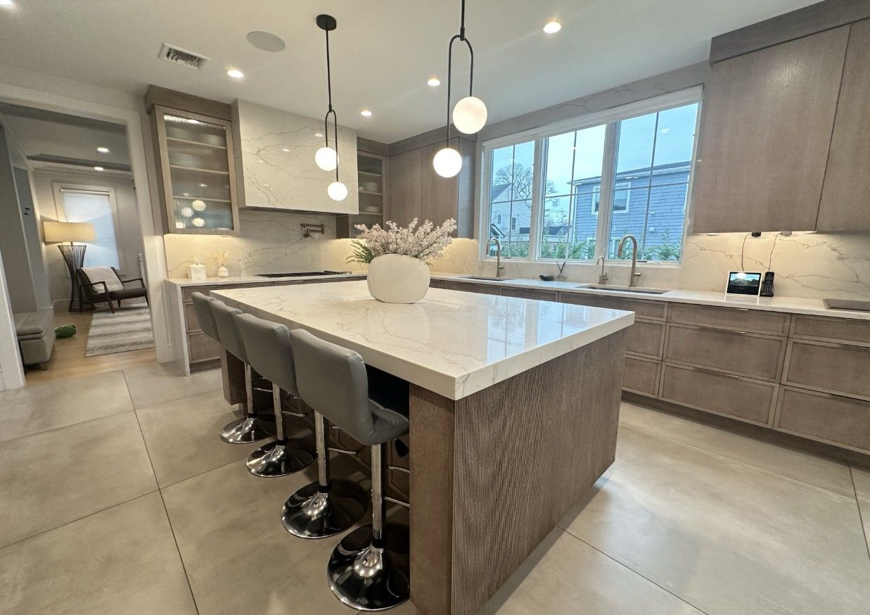 A modern, minimalist kitchen featuring a large white quartz island with three gray stools, light wood cabinets, and tile.