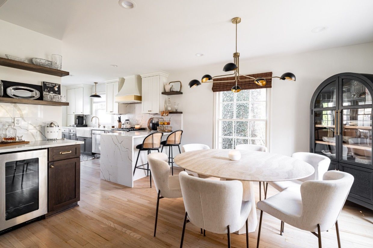A bright, modern kitchen and dining area with a marble island, white upholstered chairs, and a dark arched cabinet.