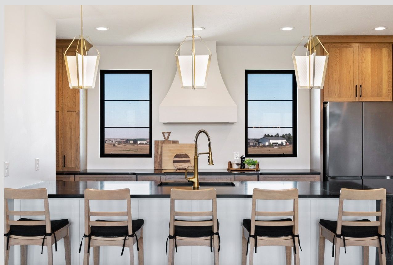 A modern kitchen features a central island with five light-wood chairs, a gold faucet, and symmetrical windows flanking a hood.