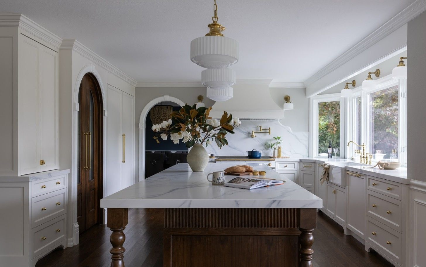 A bright, elegant kitchen featuring a large marble-topped island with wood legs and a dark wood arched doorway.