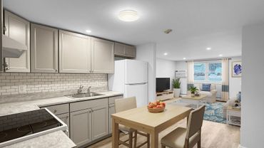 A small kitchen with light-colored cabinets and countertops, open to a living area with a window and TV.