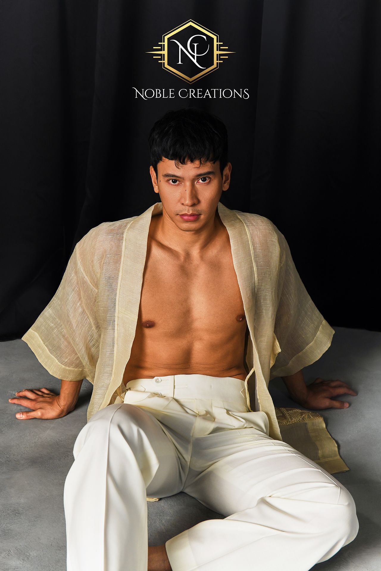 A man in an open, textured beige barong and white trousers sits on a gray floor