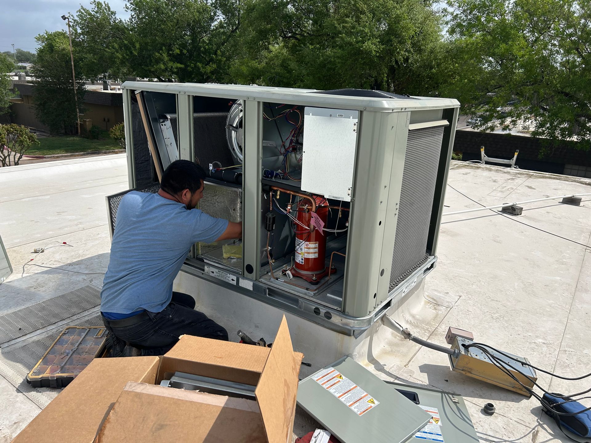 A man working on an HVAC unit on a rooftop. Gray unit, red tank, blue shirt, sunny day.