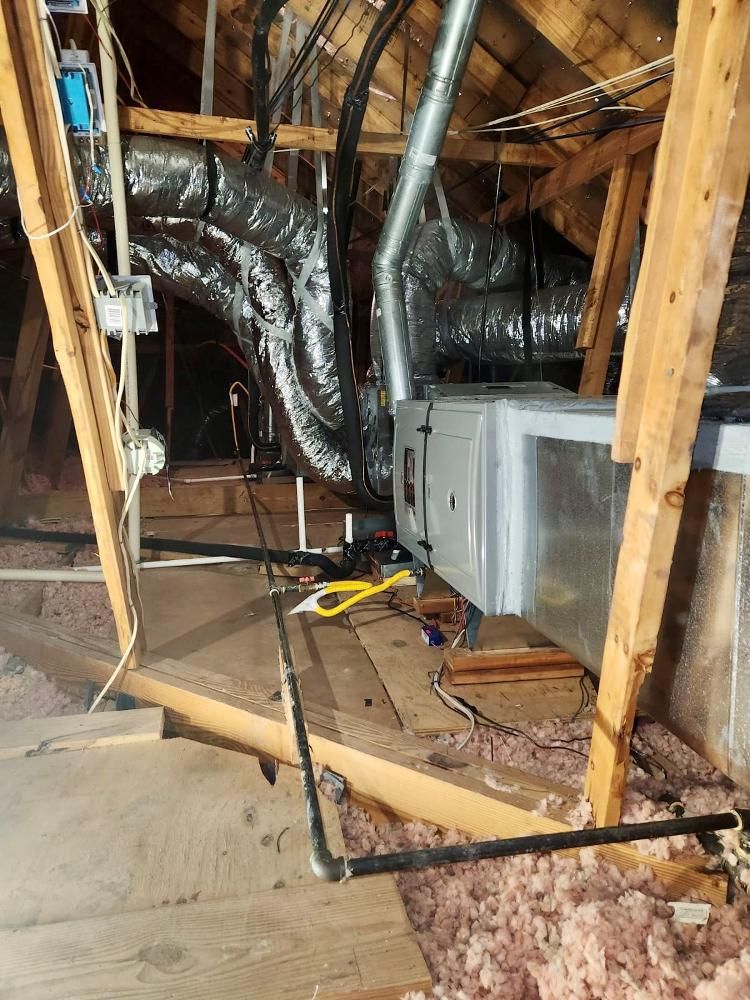 HVAC unit and ductwork in an attic. The unit is silver with black pipes. Wood framing and pink insulation are visible.