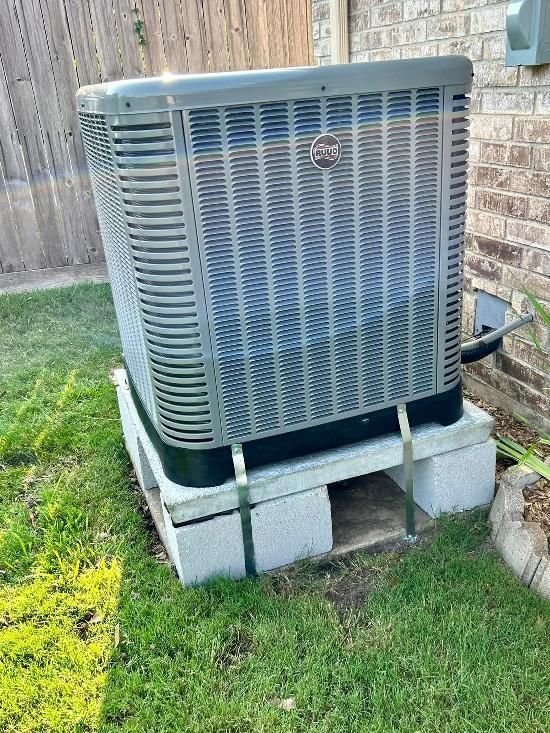 A GE air conditioning unit sits on concrete blocks in a grassy yard, next to a brick wall and wooden fence.