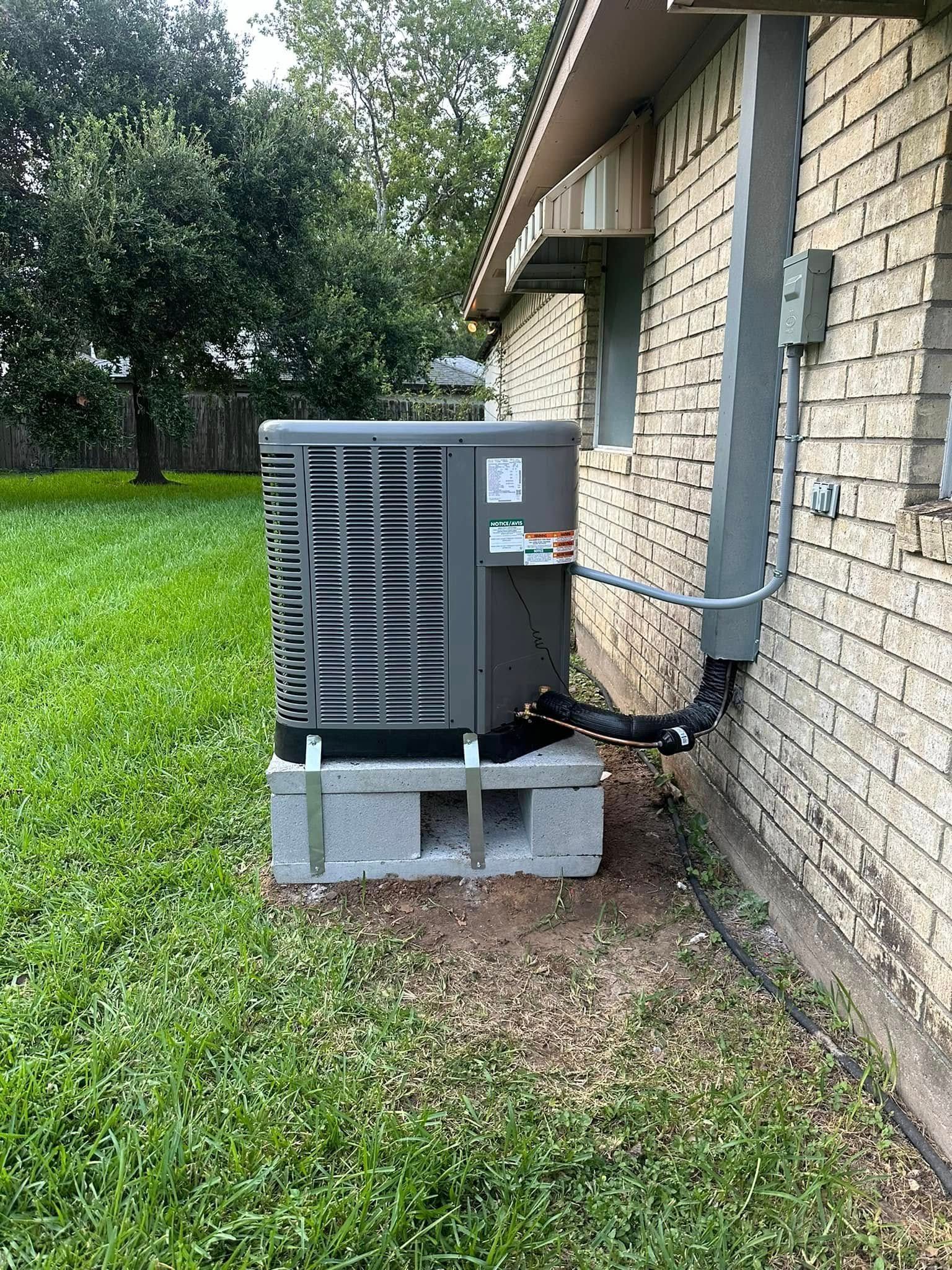 An air conditioning unit sits on cinder blocks next to a brick building. A black pipe runs from the unit towards the wall.
