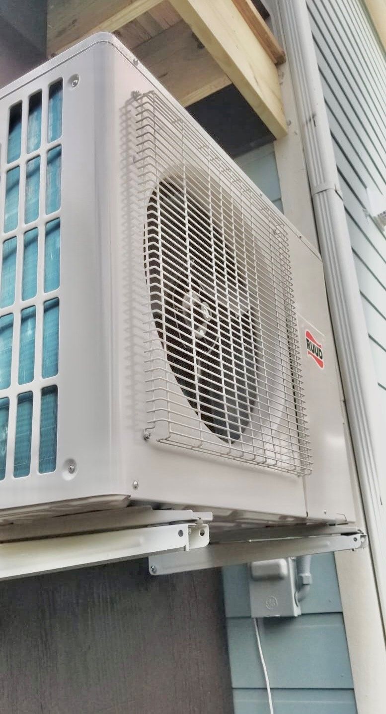 White wall-mounted air conditioner unit with a metal cage over the fan. The unit is attached to the side of a building.