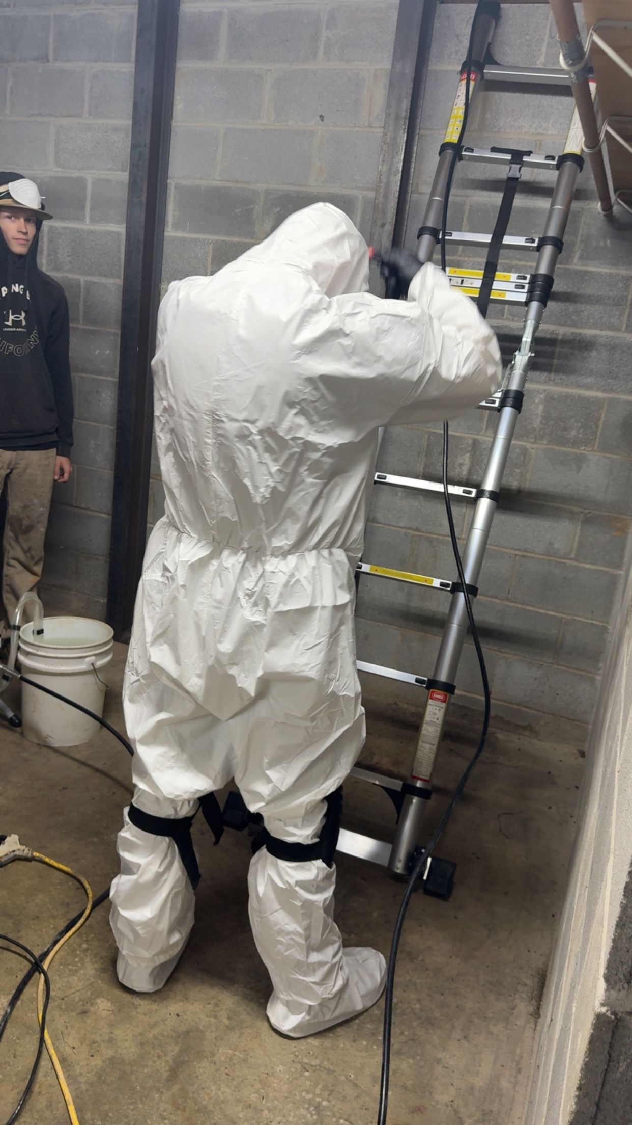 Person in white protective suit climbing a ladder in a cinderblock room, with another person standing nearby