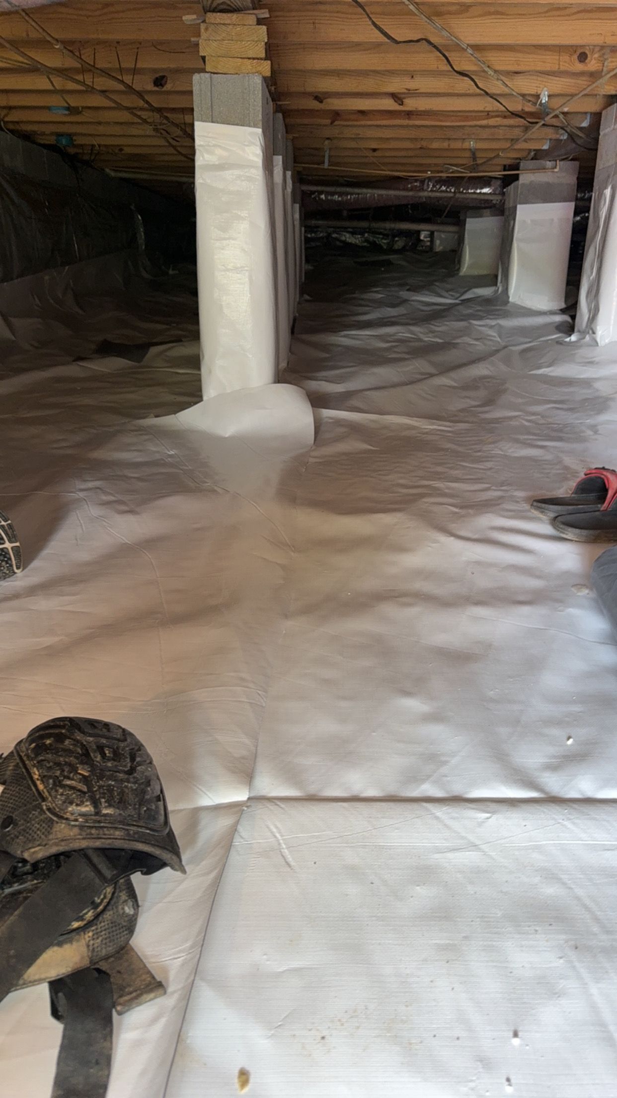 Crawlspace with white vapor barrier on floor, exposed wooden joists, and concrete support posts