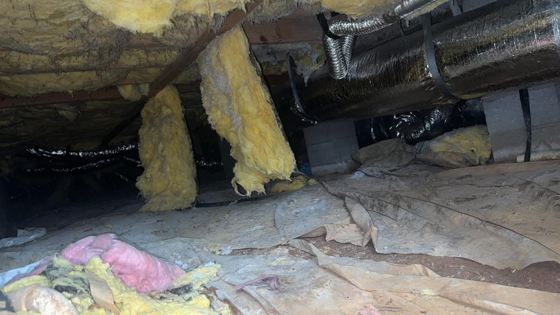 Inside a crawl space, yellow insulation hangs from joists above dirt and debris on the floor