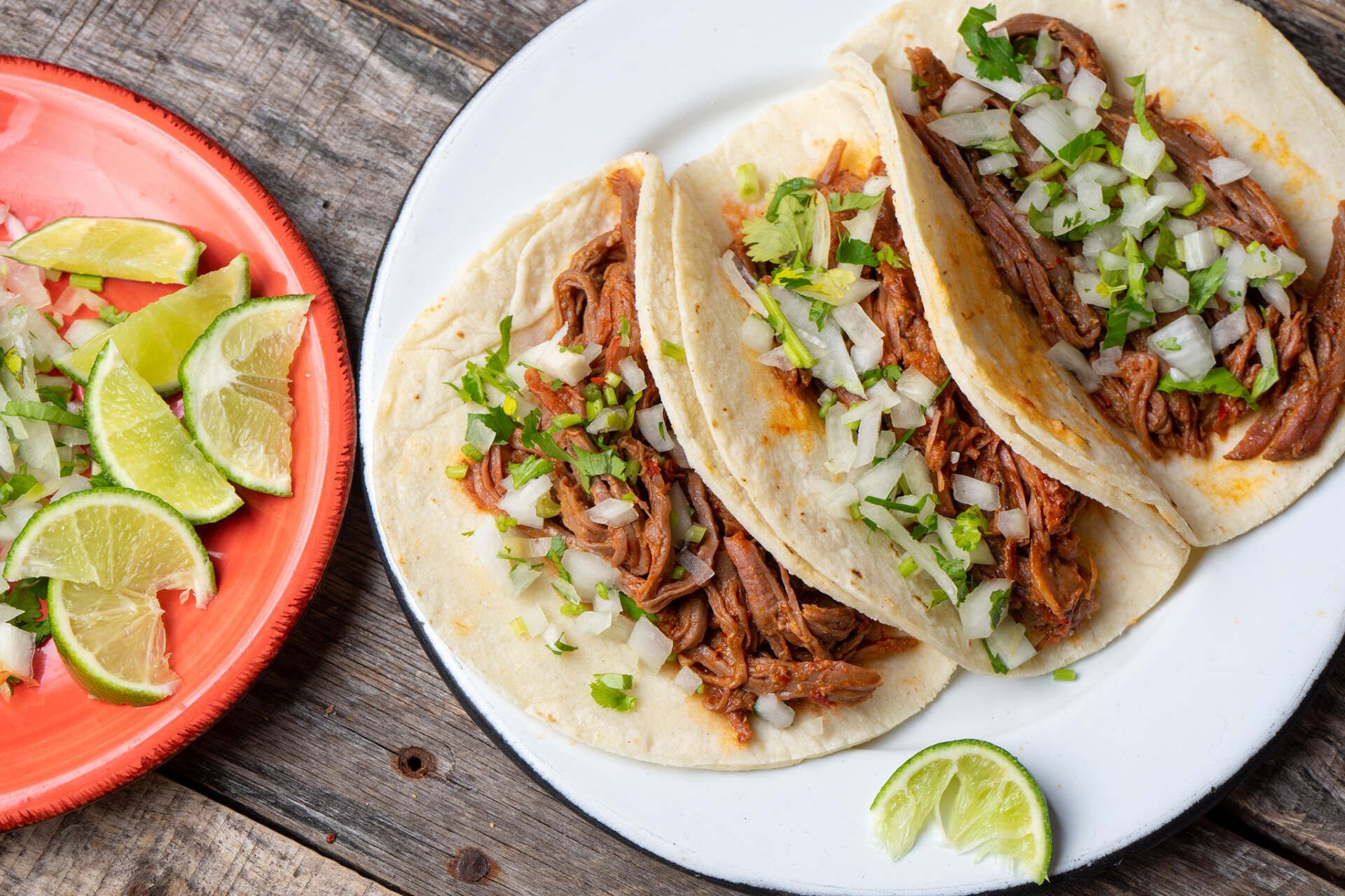 A white plate topped with three tacos next to a red plate with lime wedges.