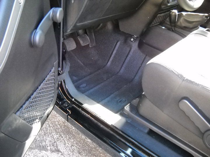 Interior view of a Jeep, showing the driver's side door, floor, pedals, and part of the seat. Black and gray tones dominate.