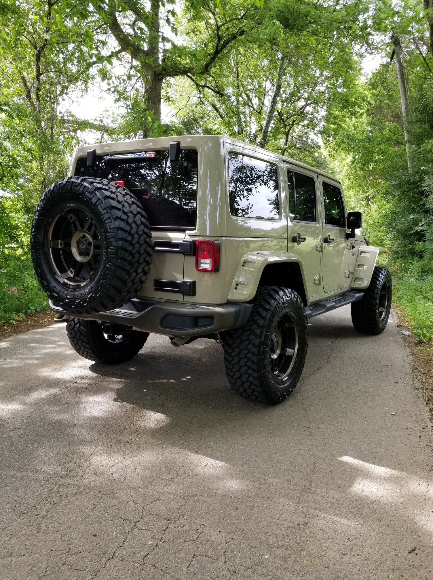 Tan Jeep Wrangler with oversized tires on a paved road, surrounded by trees. Spare tire mounted on the back.