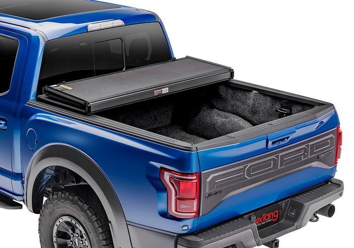 Blue Ford Raptor pickup truck with a black, folded tonneau cover. The bed is lined with dark gray material.
