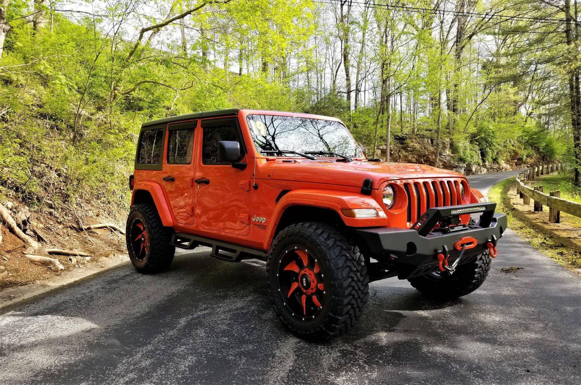 Orange Jeep Wrangler on a paved road in a wooded area. The Jeep has black and orange accents.