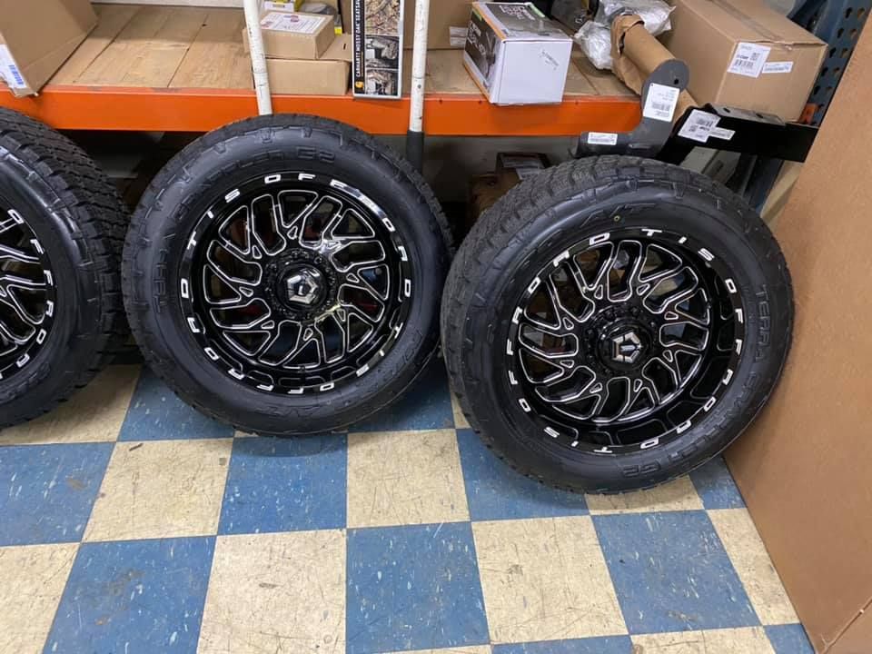 Four black tires with silver and white rims, indoors on a blue and white tiled floor, near shelves.