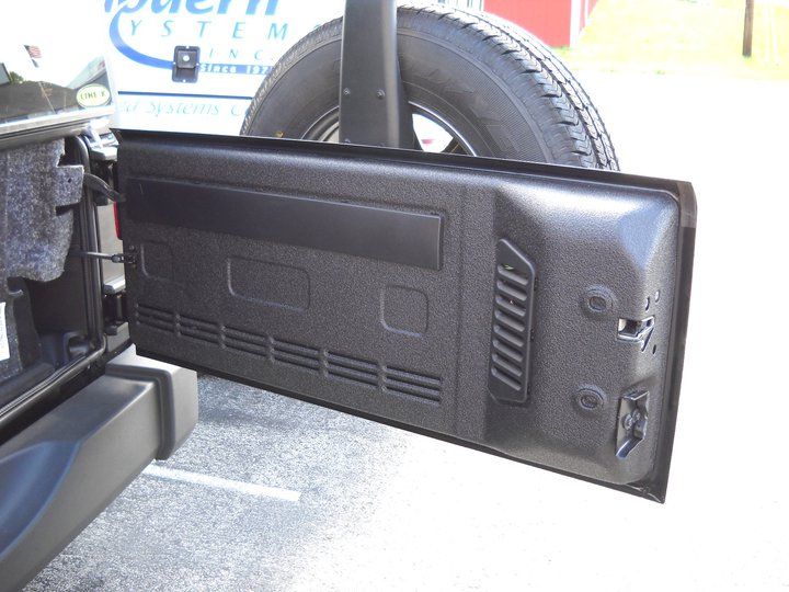 Black Jeep Wrangler rear door open, showing textured interior panel with vents and latching hardware. Spare tire is mounted behind.