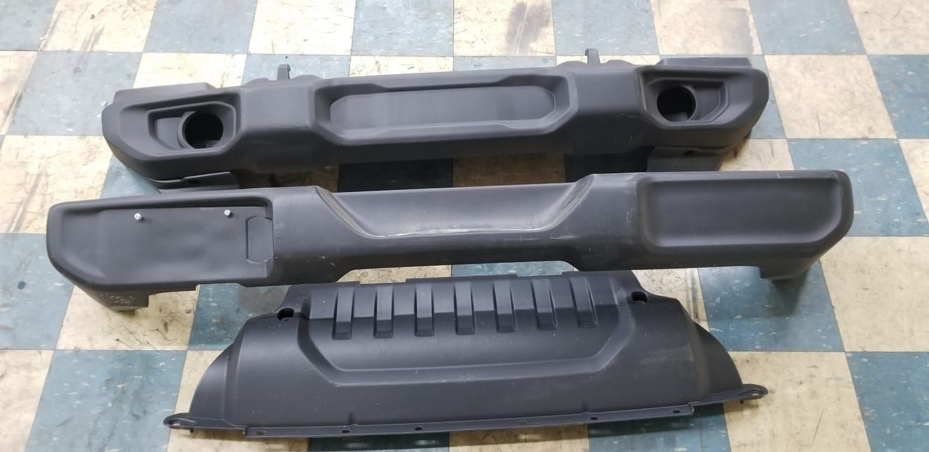 Three black car bumpers arranged on a checkered floor. The top one has two holes, the middle has a license plate mount.