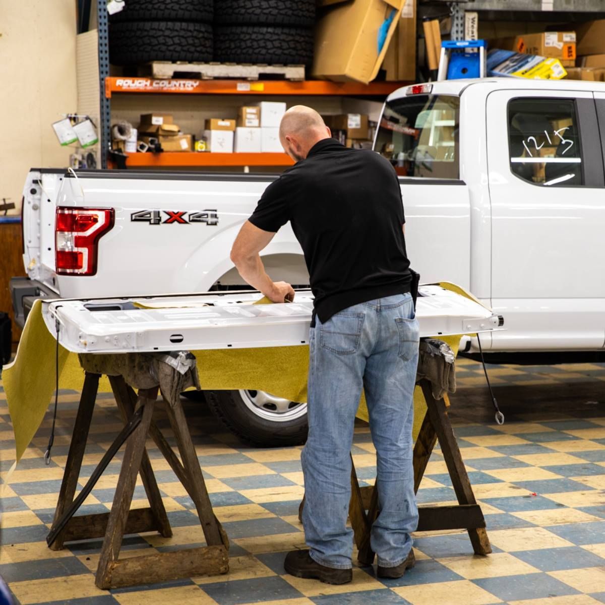 A man working on a white truck tailgate, resting on sawhorses inside a garage. He wears a black shirt and jeans.