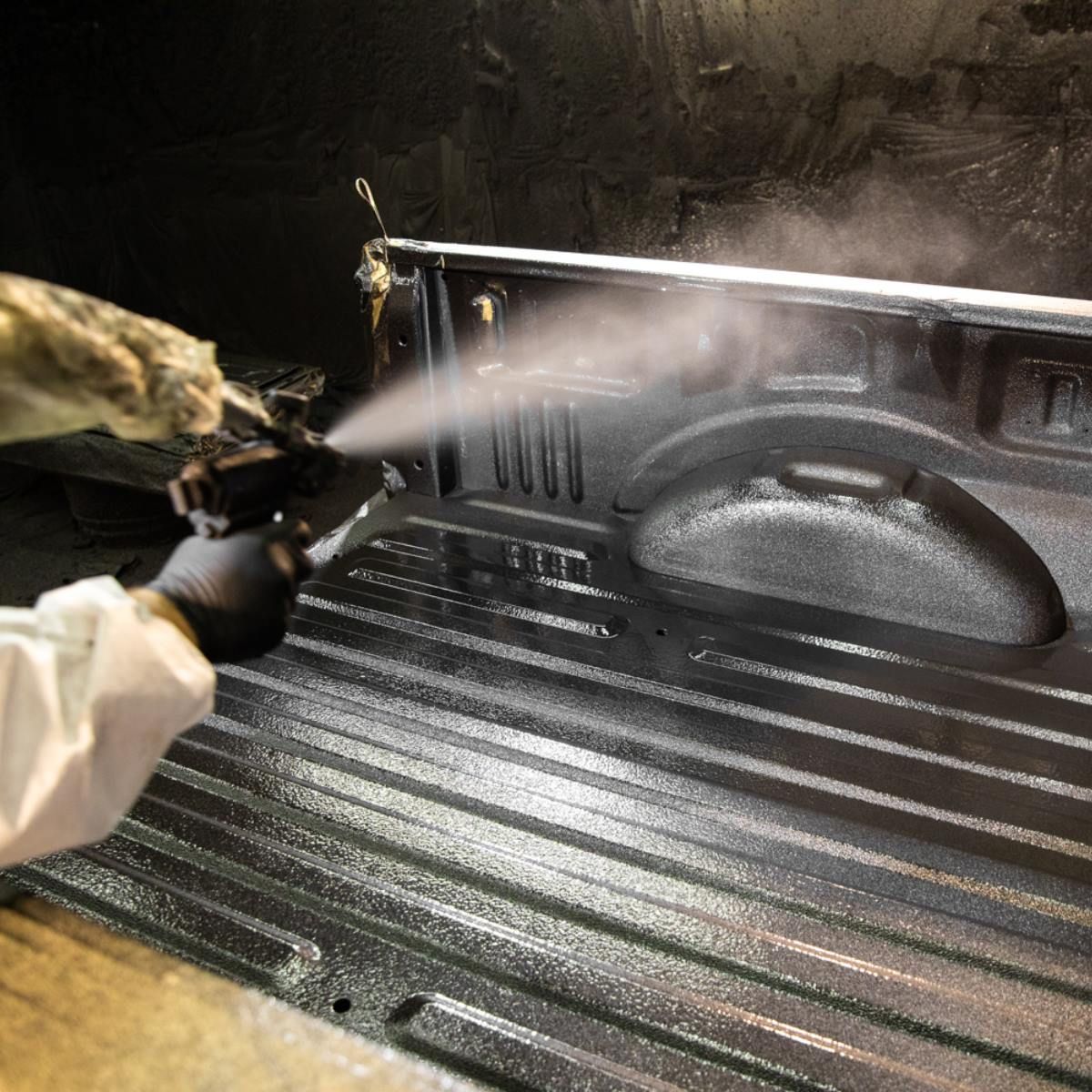 Person spraying bed liner coating inside a truck bed with a spray gun. The truck bed is black.