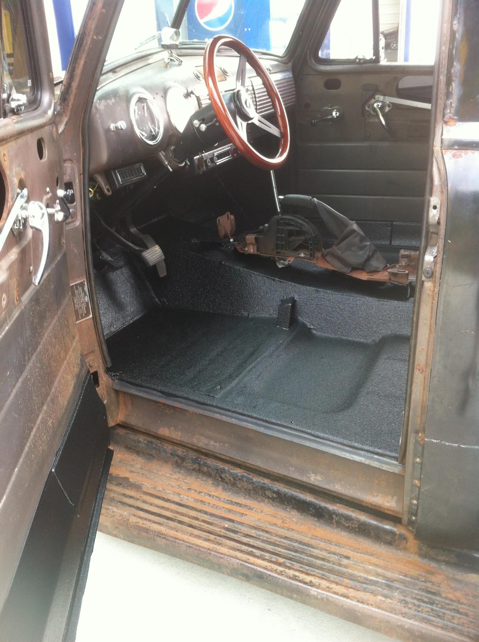 Interior of an old rusty truck with a black floor and wood steering wheel. The door is open.