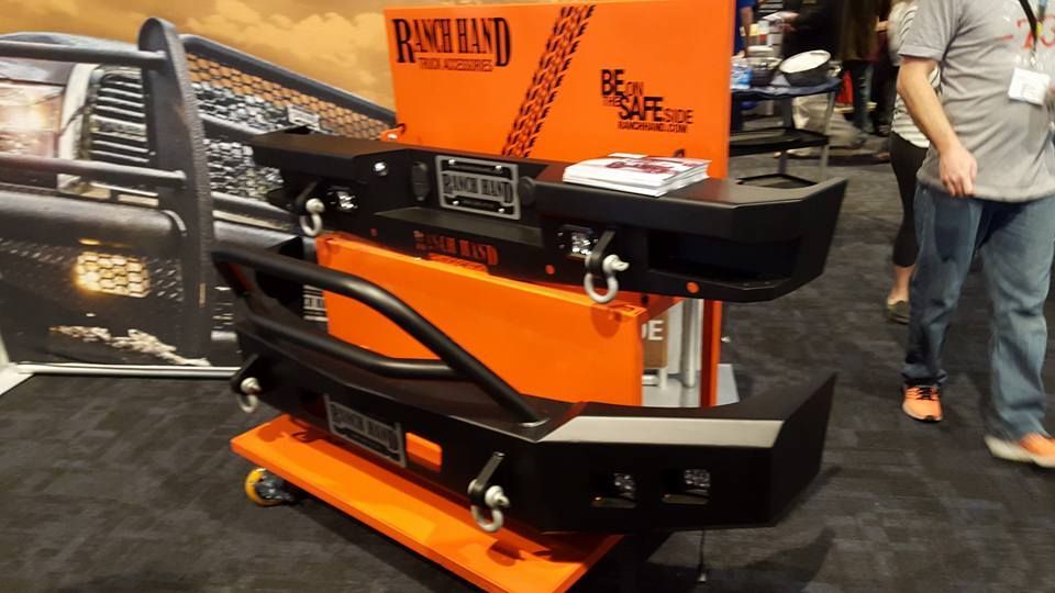 Two black truck bumpers on display at a trade show, stacked on an orange cart.  The Ranch Hand Accessories logo is visible.