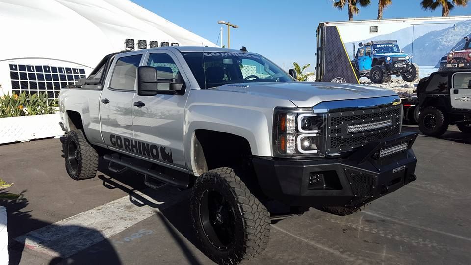 Silver lifted pickup truck with black accents, light bar, and off-road tires, parked outside under a blue sky.