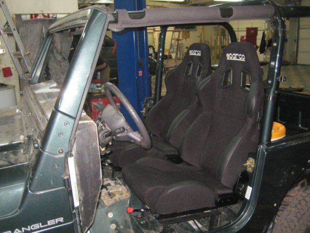 Inside a green Jeep Wrangler, two black racing seats are installed. The steering wheel is visible, and the setting appears to be a garage.