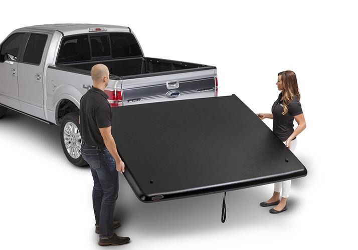 Two people installing a black truck bed cover on a silver pickup truck. The cover is being lifted towards the open bed.