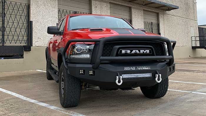 Red Ram pickup truck with black accents parked in front of a tan building. It has a black front bumper with a bull bar and a light bar.