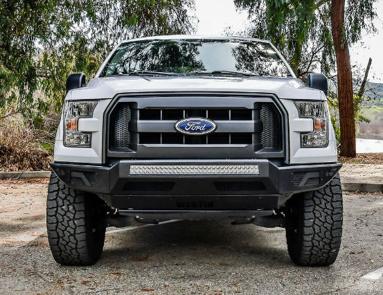 White Ford F-150 truck with black grill and light bar, parked on a gravel road in front of trees.