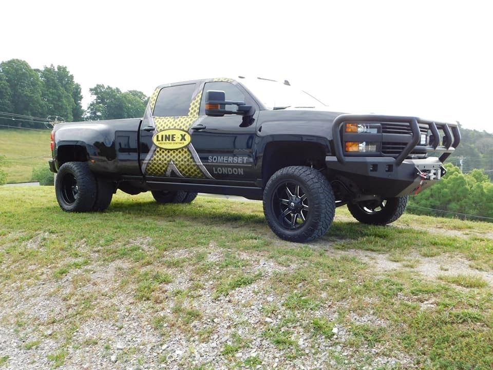 Black lifted pickup truck with Line-X logo, brush guard, and dual rear wheels parked on a grassy hill.