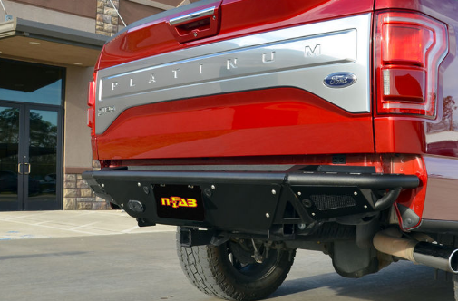 Red Ford pickup truck with a black aftermarket rear bumper. The truck is parked outside a building.