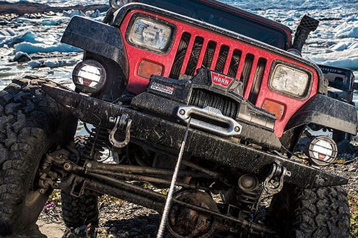 Red Jeep with a winch and large tires, covered in mud, angled against a backdrop of ice and snow.