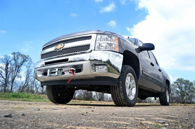 Silver Chevrolet pickup truck on a gravel road, angled upwards. Bright, sunny day with blue sky and few clouds.
