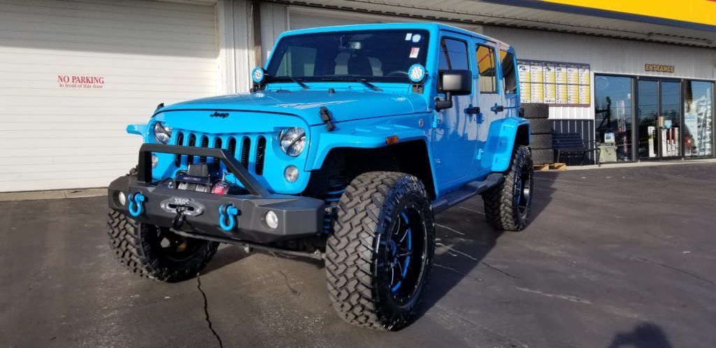 A bright blue Jeep Wrangler parked in front of a building. It has black wheels and a modified front bumper.