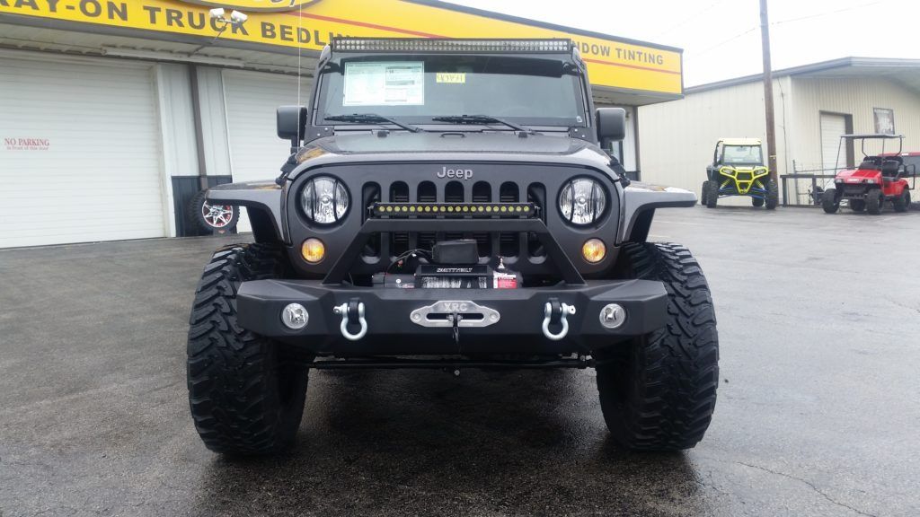 Modified black Jeep Wrangler with large tires and front winch, parked in front of a building.