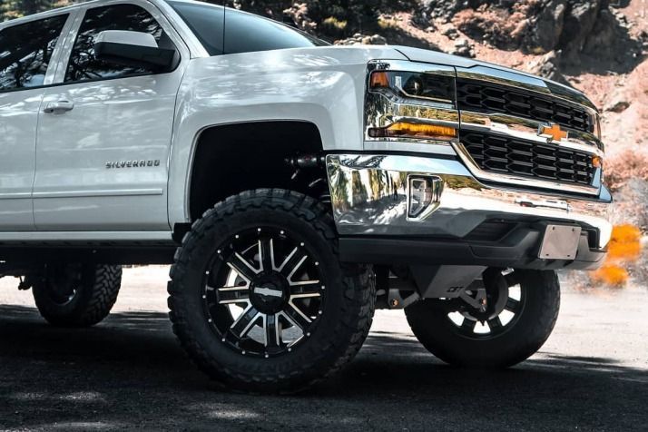 White Chevrolet Silverado pickup truck with black and chrome rims, lifted suspension, parked on a paved road near a rocky hillside.