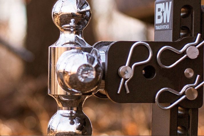 A close-up of a shiny, dual-ball trailer hitch with safety pins, set against a blurred outdoor background.