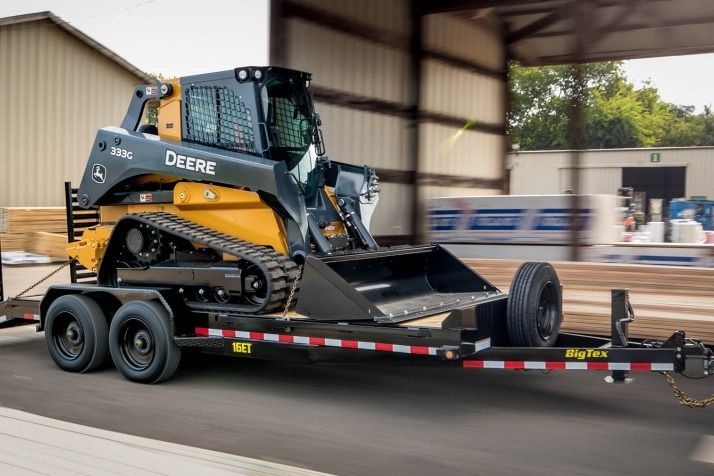 A John Deere skid steer loader on a trailer, with yellow, black, and gray colors, parked inside a building.