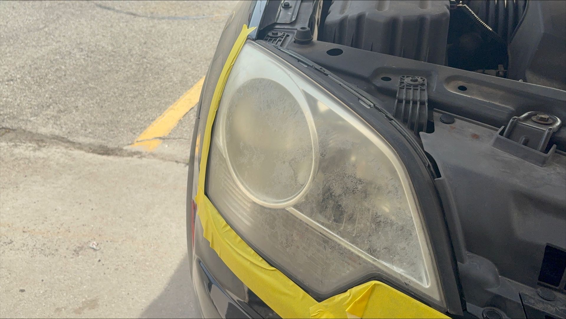 Front of a black car with a cloudy headlight covered in yellow masking tape, hood open.