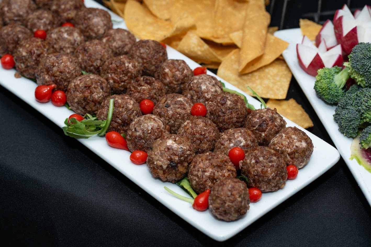 Platter of meatballs garnished with cherry tomatoes, alongside chips and broccoli on a table