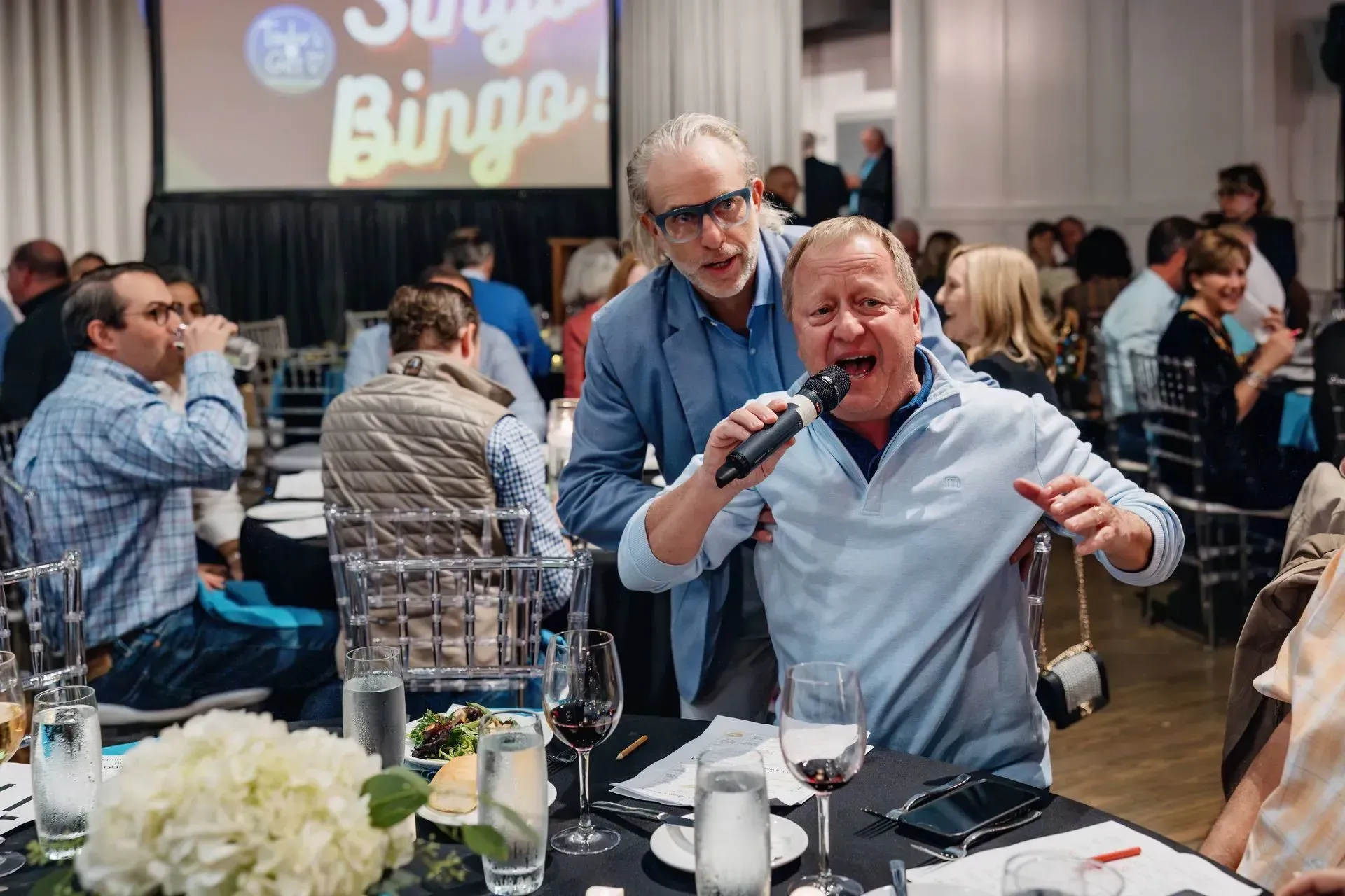Man singing into a microphone at a lively banquet, with a man standing behind him in a crowded hall